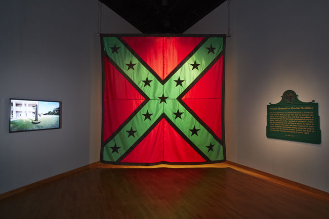 Left to right: John Sims, Freedom Memorial at Gamble Plantation, 2020. 7:47 min. video animation with sound; John Sims, AfroConfederate Flag: 12 Foot, 2020. Nylon; John Sims, Freedom Memorial Marker, 2020. Latex paint on synthetic material. All works courtesy of the artist. Installation View of Marking Monuments exhibition at USF Contemporary Art Museum. Photo: Will Lytch.